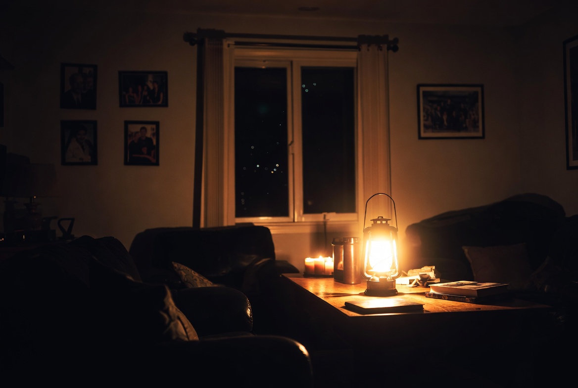 A dark living room during a power outage, lit only by a hurricane lantern and a few candles on a coffee table — leather sofas in shadow, framed family photos on the walls, night visible through the window.