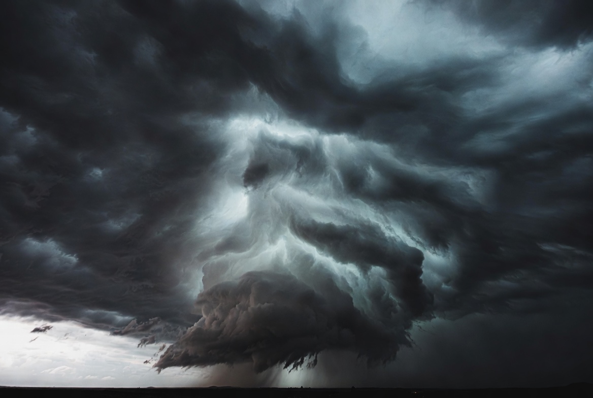 A massive dark supercell storm cloud builds over flat terrain at dusk, rain falling beneath it.